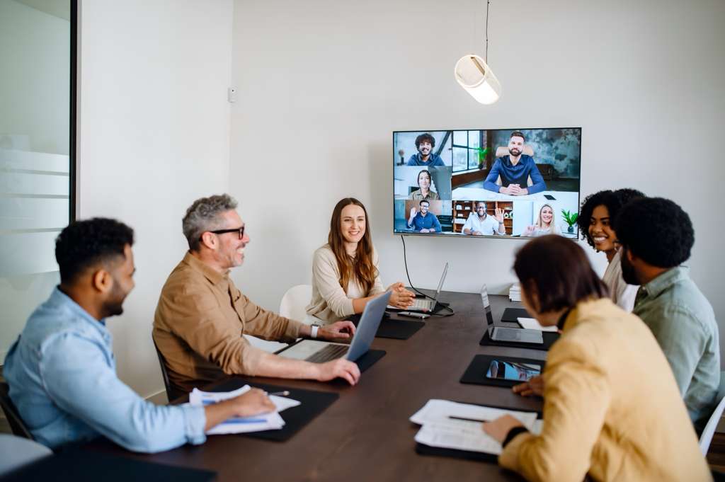 Diverse team in conference room having hybrid meeting with colleagues on video screen